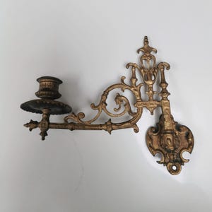 May include: Ornate, antique-style wall sconce in a gold finish. The sconce features a candle holder, decorative scrollwork, and a mounting plate. A classic piece for home decor.