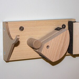 Könnte beinhalten: Eine Wandhalterung aus Holz mit zwei Haken zum Aufhängen von Gegenständen. Ein Haken ist eine einfache, gebogene Form, während der andere ein komplexeres, abgewinkeltes Design hat. Die Halterung ist aus hellem Holz gefertigt und hat eine dunkelbraune Rückseite.