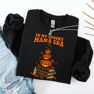 May include: A black crewneck sweatshirt featuring the orange text IN MY SPOOKY MAMA ERA. The graphic includes a stack of three carved jack-o'-lanterns perched on a pile of three old books, with a black crow on top, a lit candle, and autumn leaves.