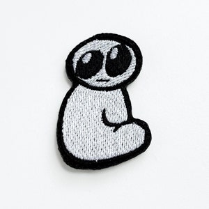 Könnte beinhalten: Ein gestickter Aufnäher mit einer Cartoonfigur mit großen schwarzen Augen und einem einfachen, abgerundeten Körper. Die Figur sitzt mit gekreuzten Beinen.