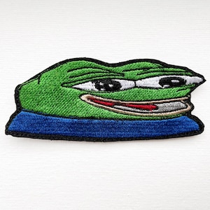 Könnte beinhalten: Grüner gestickter Aufnäher eines lächelnden Frosch-Memes mit einem blauen Kragen. Der Frosch hat ein breites Lächeln mit einer roten Zunge und weißen Augen.