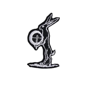 May include: Black and white embroidered patch featuring a rabbit holding a circular object with a cross design. The patch has a detailed outline and is likely for clothing or accessories.