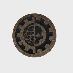 Könnte beinhalten: Runder Aufnäher mit geteiltem Design: ein Totenkopf und eine mechanische Kreatur in einem Zahnrad. Der Aufnäher hat einen beigen Hintergrund mit schwarzen Details. Ideal für Jacken oder Taschen.