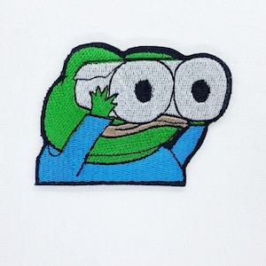 May include: Embroidered patch featuring a green frog-like character with large, white eyes and a blue shirt. The character is holding its hands up to its eyes, as if looking through binoculars. The patch has a black border and is on a white background.