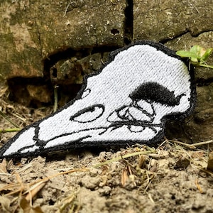 Könnte beinhalten: Weißer gestickter Aufnäher mit einem Vogelschädel mit schwarzen Details. Der Schädel ist nach links gerichtet und hat einen langen Schnabel.
