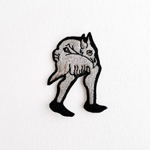 May include: Embroidered patch with a surreal design. It depicts a figure with a human face and legs, carrying a creature. The patch is mainly grey with black outlining and black feet. The design is detailed and artistic.