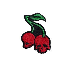 May include: Two red cherries with skull faces and green stems. The cherries are embroidered on a black fabric patch.