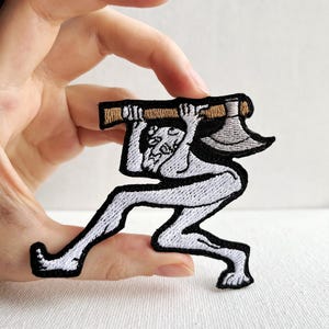 Op de afbeelding: Witte geborduurde patch van een figuur die een bijl boven zijn hoofd houdt. De figuur rent en de bijl is bruin met een zilveren blad.