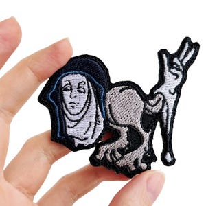Puede incluir: Parche bordado para planchar con una ilustración en blanco y negro de una monja con una calavera y huesos cruzados. La monja lleva una túnica negra con un velo blanco y un ribete azul. La calavera lleva un sombrero negro.