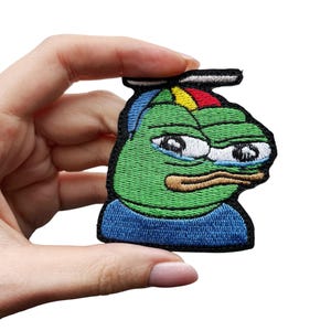 Puede incluir: Parche bordado con una rana verde de dibujos animados con una expresión triste. La rana lleva una camisa azul y un sombrero con detalles rojos, amarillos y azules. El parche está sobre un fondo blanco.