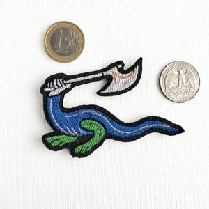 Op de afbeelding: Een geborduurde patch met een blauwe zeeslang die een bijl vasthoudt. De slang heeft groene schubben en wordt afgebeeld in een dynamische pose.