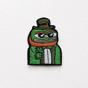 Puede incluir: Rana verde con un sombrero verde con una hebilla dorada, un traje verde y una barba naranja. La rana sostiene un trébol de cuatro hojas en su mano.