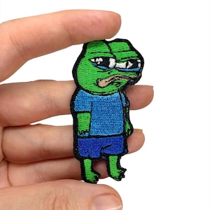 Puede incluir: Parche verde bordado para planchar de una rana triste con una camisa azul y pantalones cortos azules.