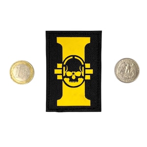 May include: A rectangular black fabric patch featuring a large yellow capital letter I with a stylized skull icon centered within the letter. The patch is positioned between a one euro coin and a United States quarter coin for scale reference.