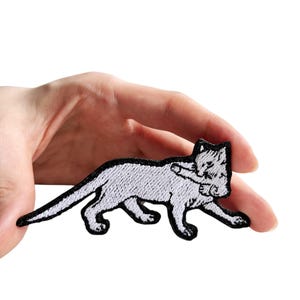 May include: Embroidered patch featuring a stylized white cat with black outline and details. The cat is depicted in a walking pose, with its tail extended. The patch is held in a hand against a white background.
