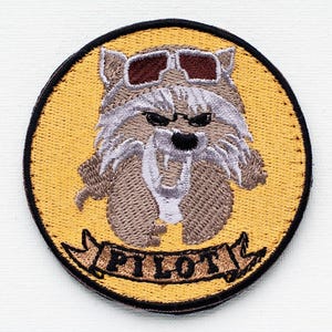 Op de afbeelding: Gestikte patch met een cartoonwolf die een zonnebril draagt en een banner met de tekst "PILOT". De wolf is bruin en wit op een gele achtergrond.