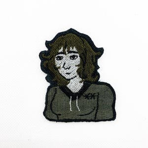 May include: Embroidered patch featuring a stylized illustration of a person with brown hair and a green hoodie. The patch has a black border and the person's face is white. The hoodie has the letters "XX" on the front.