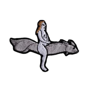 May include: Embroidered patch featuring a nude figure riding a stylized creature. The figure has blonde hair and is seated on a gray, textured animal with a black outline. The patch is designed for application on clothing or accessories.