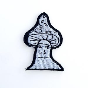 以下が含まれることがあります： 顔と黒い縁取りのある水色のキノコの刺繍パッチ。キノコの傘には白い点があります。このパッチは、衣類やアクセサリー用です。