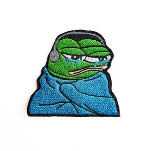 Puede incluir: Parche bordado con una rana verde con auriculares, llorando. La rana lleva una prenda azul. El parche tiene un borde negro y está sobre un fondo blanco.