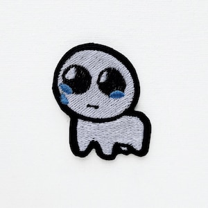 Könnte beinhalten: Gestickter Aufnäher mit einem kleinen, grauen, außerirdischen Wesen mit großen schwarzen Augen und blauen Tränen. Der Aufnäher hat einen schwarzen Rand und ein schlichtes Design, ideal für Kleidung oder Accessoires.