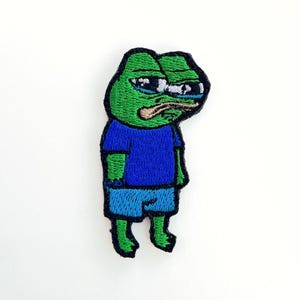 Puede incluir: Parche bordado de un meme de rana verde con una camisa azul y pantalones cortos azules. La rana tiene una expresión triste con una sola lágrima que le cae del ojo.
