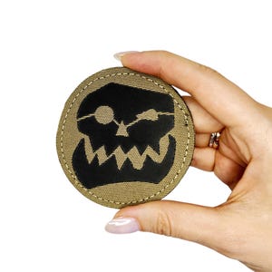 Puede incluir: Un parche circular de tela color canela con un gráfico bordado en negro de una calavera estilizada con una boca dentada y un parche en el ojo. El parche tiene un borde cosido alrededor y un acabado de tejido texturizado en el fondo canela.