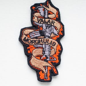 Puede incluir: Un parche bordado naranja y negro con un rifle estilizado y las palabras "Valar Morghulis" en una pancarta beige.