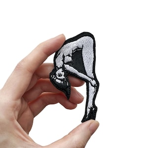 May include: Embroidered patch in black and white, depicting a stylized figure in a contorted pose. The patch has a black border and is held against a white background. The figure is bent over.