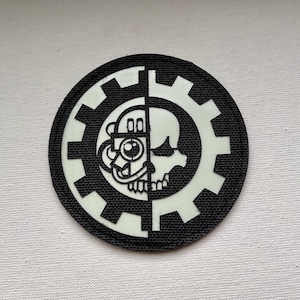 Puede incluir: Parche redondo en blanco y negro con diseño de engranaje. El diseño está dividido por la mitad, con una cara mecánica en un lado y una calavera en el otro. El engranaje y la cara/calavera son blancos, sobre fondo negro.