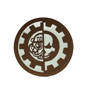 May include: A round, brown patch featuring a gear design with a half-skull and half-mechanical face in white. The patch has a textured appearance and is set against a white background. The design is a blend of mechanical and human elements.