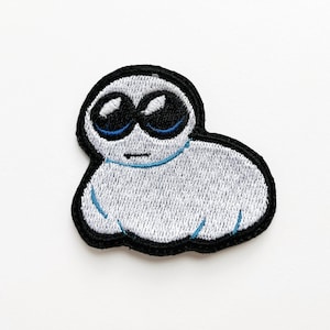 Könnte beinhalten: Gestickter Bügel-Patch mit einer weißen, cartoonartigen Kreatur mit großen, schwarzen Augen und blauen Pupillen. Die Kreatur hat einen runden Körper und einen einfachen, lächelnden Mund.