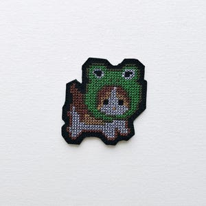 Puede incluir: Un parche de punto de cruz que representa un perro con un disfraz de rana verde. El parche tiene un borde negro y está hecho con hilo marrón, blanco y verde. La cara del perro es visible y el disfraz de rana tiene ojos grandes.