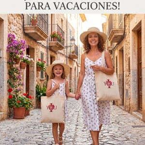 May include: A mother and child walk down a cobblestone street, both wearing straw hats and holding matching tote bags. The bags are beige with a red floral design and the word "YOU". The text at the top says "A perfect duo for vacations!"
