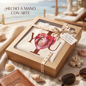 May include: A gift box with a canvas tote bag that says "YOU" with a heart design. The box is tied with a ribbon and a tag that says "A Perfect Gift for your Vacation". Seashells, sunglasses, and a passport are also in the image.