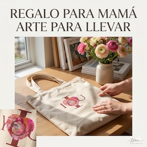 May include: A beige tote bag with the letters "I U" in a burgundy color, with a floral design in the center. The bag is on a wooden surface, with a vase of flowers and books in the background. The text "REGALO PARA MAMÁ ARTE PARA LLEVAR" is at the top.