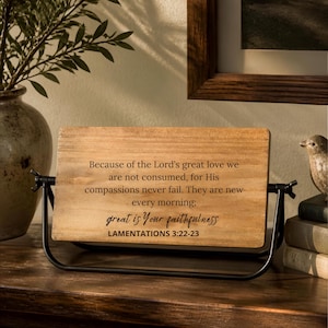 May include: A rectangular wooden sign with a natural light brown finish, held by a black metal swivel frame. The sign features black printed text that reads: Because of the Lord's great love we are not consumed, for His compassions never fail. They are new every morning; great is Your faithfulness LAMENTATIONS 3:22-23.
