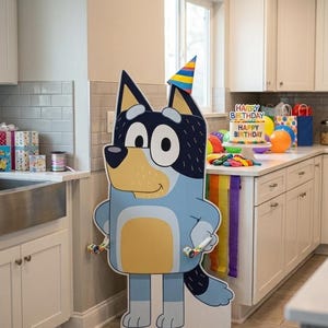 May include: A large, flat, cardboard cutout of a blue and tan cartoon dog character wearing a colorful striped party hat. The character has large white eyes, a black nose, and is holding two party blowers in its hands, standing upright.
