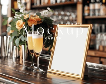 May include: A gold picture frame with a white background sits on a wooden bar counter. Two champagne flutes filled with a yellow beverage are in front of the frame. A bouquet of flowers is behind the frame.