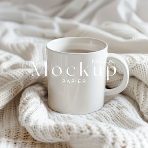 以下が含まれることがあります： 白いニットの毛布の上に置かれた、薄い茶色の液体が入った白いセラミック製のマグカップ。マグカップには「Mockup Papier PSD JPG」という文字が印刷されています。