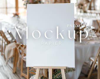 May include: A white rectangular sign with the word "Mockup" and "Papier" in a light font, displayed on a wooden easel. The sign is in a bright, airy room with tables and chairs, suggesting a wedding or event setting.