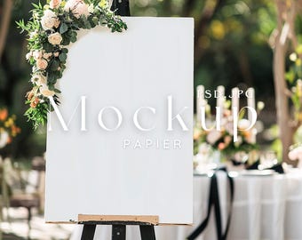 May include: White easel with a blank sign, decorated with a floral garland. The sign is set up outdoors at a wedding reception with a white tablecloth and black and white decorations.