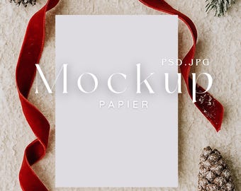 May include: A white rectangular paper mockup with the word "Mockup" and "Papier" printed on it. A red velvet ribbon and pine cones are arranged around the paper on a textured surface. The image has a festive, holiday theme.