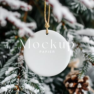 May include: White ceramic ornament with a gold string hanging from a snowy pine tree branch. The ornament has the text "Mockup Papier JPG" printed on it.