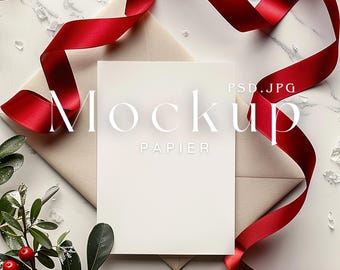 May include: A white card with the word "Mockup" and "Papier" printed on it, placed on a beige envelope. A red ribbon is draped around the card and envelope. Green leaves and red berries are in the lower left corner.