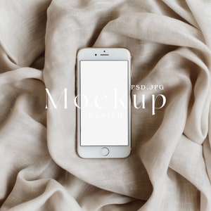May include: A white smartphone with a blank screen, featuring a gold-colored frame, rests on a beige fabric background. The word "Mockup" is displayed in a delicate font, along with the word "Papier". The image is a PSD.JPG file.