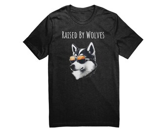 May include: A dark grey t-shirt with the text "RAISED BY WOLVES" printed in white. A cartoon husky wearing sunglasses is centered on the shirt.