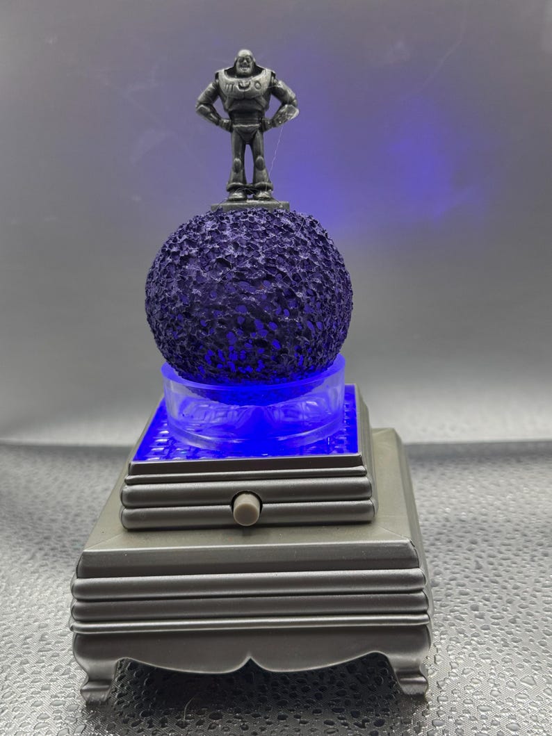 Puede incluir: Una figurita musical de plata de Buzz Lightyear de Toy Story de pie sobre una esfera p&uacute;rpura texturizada. La esfera est&aacute; sobre una base plateada con una luz azul que ilumina la esfera. La base tiene un bot&oacute;n para encender y apagar la luz.