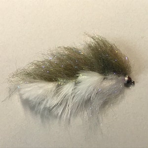 May include: A handmade fishing fly with a layered design. The fly features a combination of white and olive-green materials, with shimmering accents. The fly is designed for attracting fish and is a popular item for anglers.