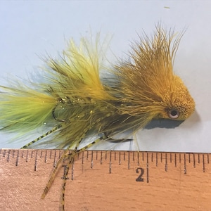 May include: A yellow and green fly fishing lure with a fuzzy body and a black eye. The lure is approximately 2.5 cm long and is sitting on a ruler.